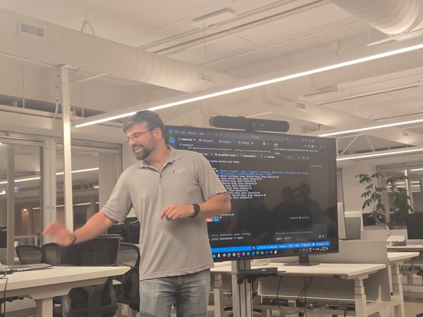A man wearing glasses and a Microsoft polo shirt is giving a technical presentation or demo in a modern office setting, standing next to a large monitor displaying code and terminal output.