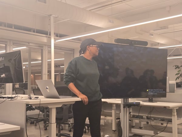 A person stands next to a large monitor displaying a "No signal" error message in a modern office, resting their arm on a standing desk with an open laptop, suggesting a presentation setup.