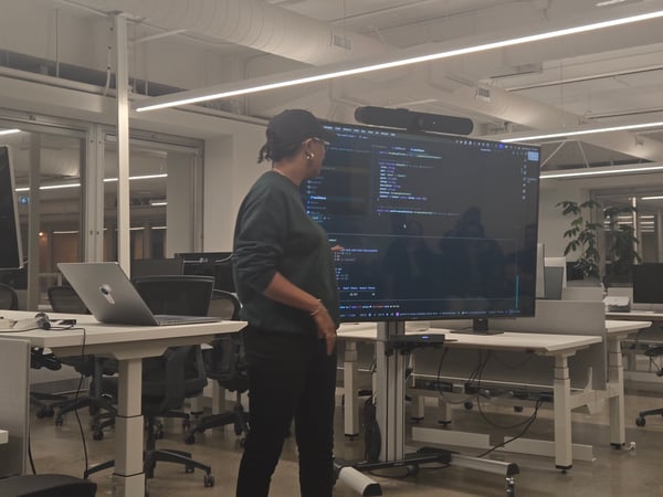 A person stands in a modern, brightly lit office space, giving a presentation or review of code displayed on a large, dark-mode monitor.