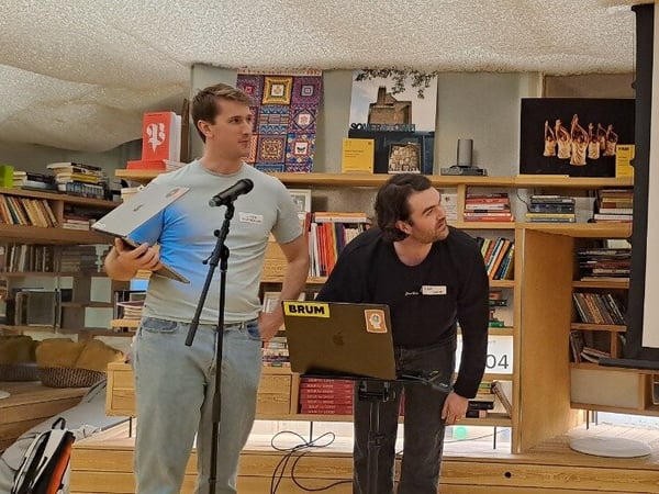 A photograph capturing two men giving a technical presentation indoors, standing in front of a projection screen displaying a dark-mode software interface and a wall of bookshelves.