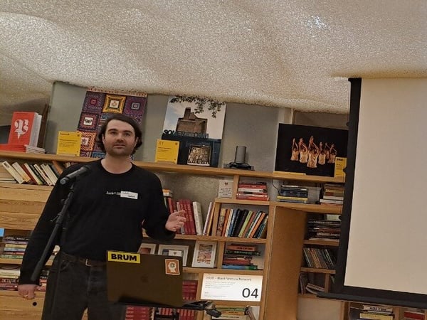 A man is giving a presentation indoors, standing next to a microphone and a laptop stand, while a large projection screen displays the name "Jack" and a list of steps related to finding a job. The background is dominated by wooden bookshelves filled with books.