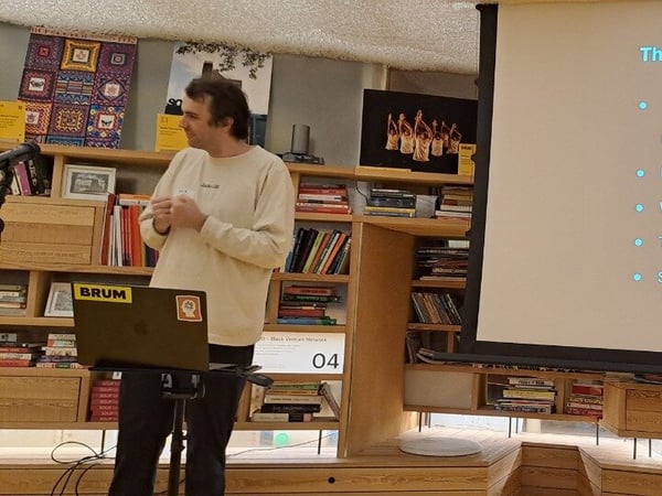 A man is giving a technical presentation in an indoor setting characterized by wooden bookshelves, standing next to a laptop while a large screen displays a bulleted slide titled "The Internal Analytics Bottleneck."