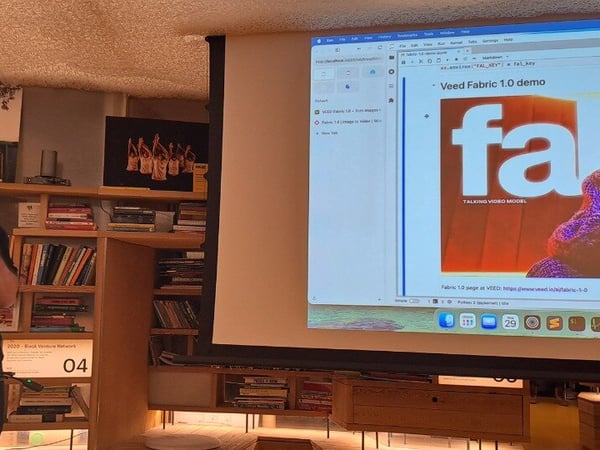 A man is giving a presentation or demo, holding a microphone in a dimly lit room characterized by wooden bookshelves, while a large screen displays a web browser showing the "Veed Fabric 1.0 demo."