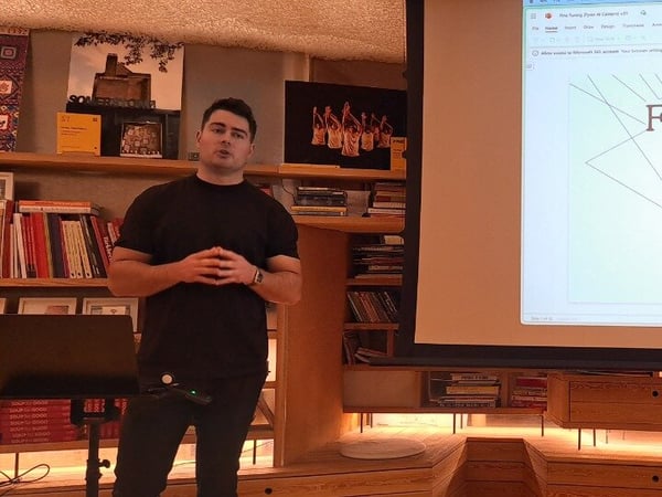 A photograph showing a man in a black shirt giving a presentation indoors, standing next to a large projection screen displaying a slide titled 'Fyxer.ai FINE TUNING FOR START-UPS'.