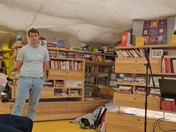 A candid photograph capturing three men participating in a discussion or presentation inside a unique, brightly lit indoor space characterized by extensive wooden bookshelves and a textured ceiling.