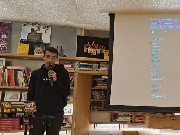 A man wearing a black hoodie and glasses is giving a technology presentation indoors, holding a microphone in front of a large projection screen that displays a web application interface titled, "Turn ideas into workflows."
