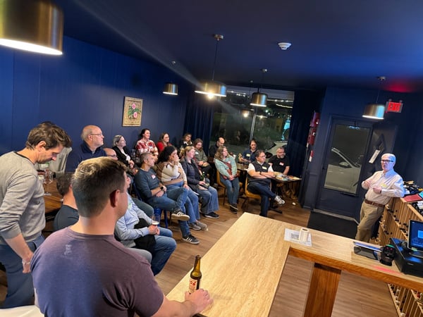 A photograph capturing an indoor social or professional gathering where an older man is giving a presentation or talk to an audience seated in a room with dark blue walls and modern lighting.