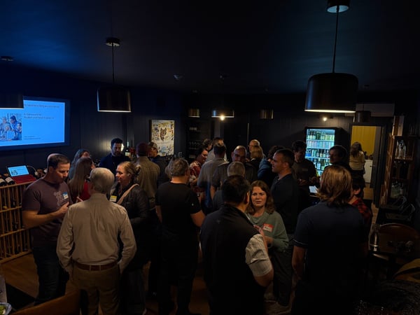 A candid photograph captures a large group of people mingling and networking in a dimly lit indoor venue, which appears to be a bar or event space with dark walls, wine racks, and a visible presentation screen on the left.