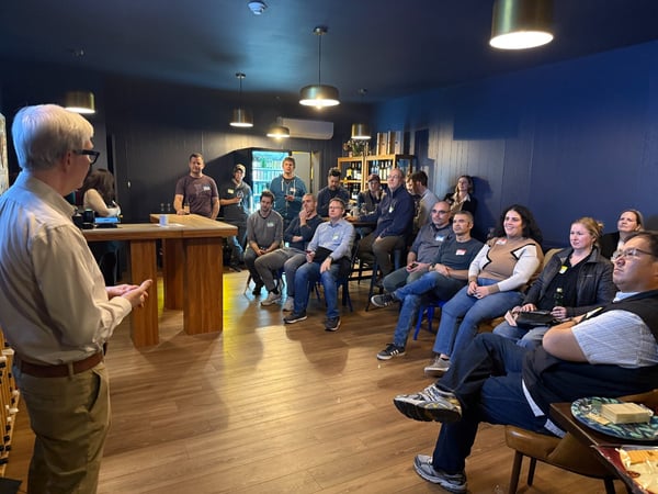 A man addresses a seated and standing audience during an indoor gathering or presentation in a dimly lit room featuring dark blue walls and wooden flooring.