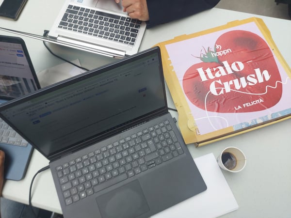An overhead photograph captures a busy desk scene featuring three open laptops, a cardboard food box with a large red tomato design and 'Italo Crush' branding, and a small cup of coffee.