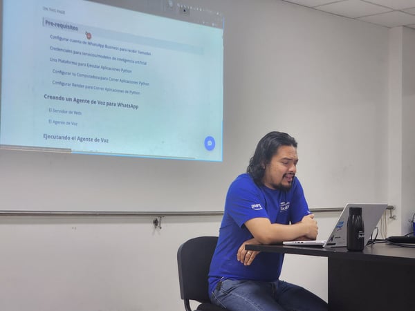 A man wearing a blue AWS Community Builder t-shirt is seated at a dark table, leaning over his open laptop during a technical presentation or workshop.