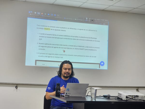 A man wearing a blue shirt is seated at a table, looking down at a laptop, while a technical presentation written in Spanish is projected onto the white wall behind him.
