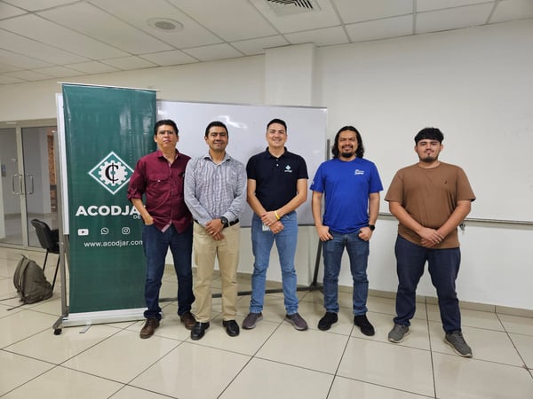 A group photograph shows five men standing side-by-side indoors, posing in front of a white wall or whiteboard and a tall green promotional banner for 'ACODJAR'.