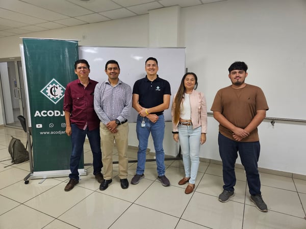 A group of five people, consisting of four men and one woman, stand side-by-side indoors, posing for a photo in front of a white wall and a dark green promotional banner.