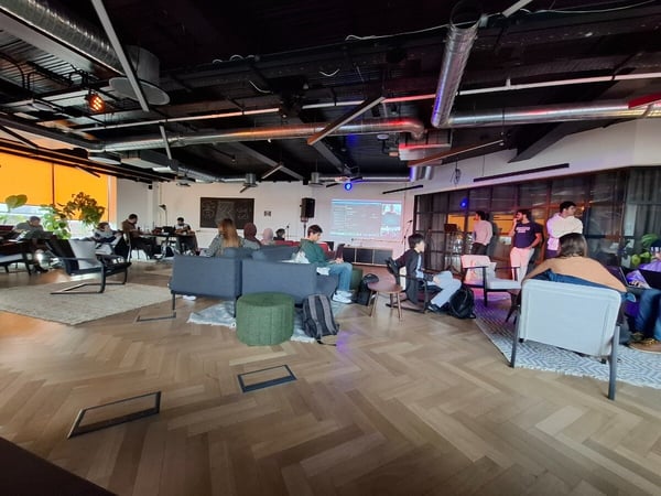 A candid photograph captures a large group of people gathered in a modern, industrial-style indoor space, featuring exposed ductwork, herringbone wood floors, and casual seating arrangements, with a screen displaying content projected onto a wall.