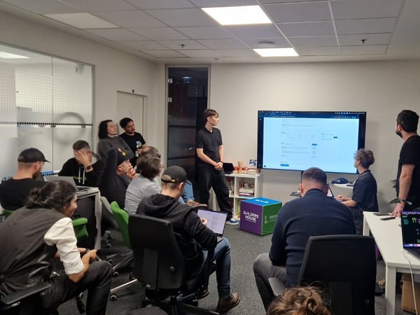 A photograph capturing an indoor meeting or presentation where a speaker stands next to a large monitor displaying application data, while approximately a dozen attendees are seated, many using laptops.