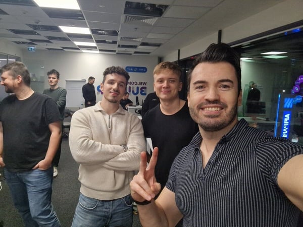 A group of men poses for a selfie inside a brightly lit modern office or meeting room, with the man closest to the camera smiling and making a peace sign.