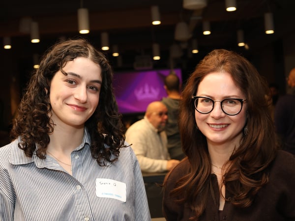 Two women are smiling at the camera during an indoor evening event with other attendees and a purple screen in the background.