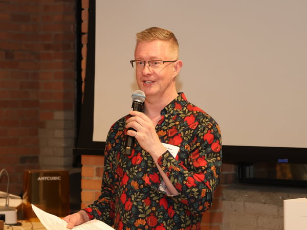 A man wearing a vibrant pomegranate-patterned shirt and glasses is speaking into a microphone during a presentation in front of a projection screen.