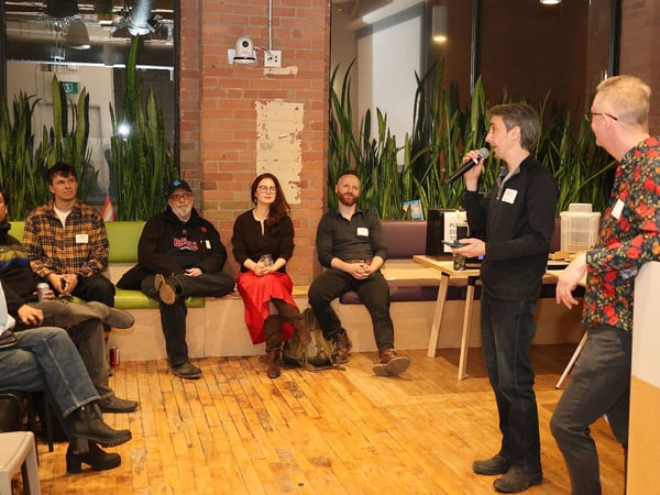 A man speaks into a microphone during a presentation or social gathering in an indoor space with brick walls and wooden floors, while several people sit and listen.