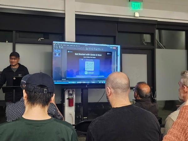A photograph taken indoors shows a technical presentation in progress, with a speaker standing next to a large screen displaying a slide titled 'Get Started with Qwen & Wan' and featuring a QR code, while several audience members are visible from behind.