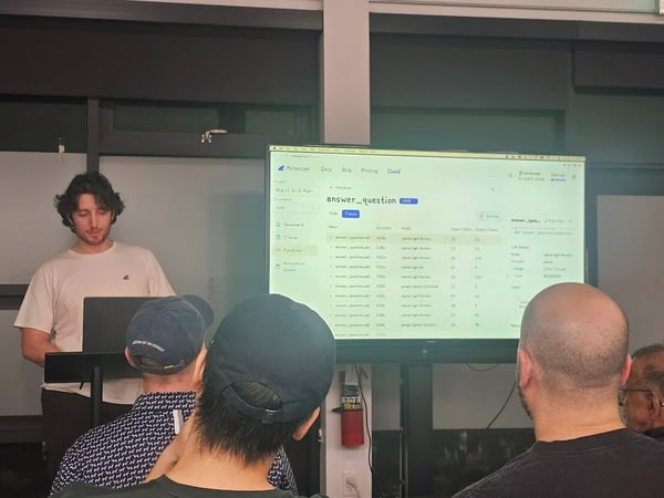 A young man stands at a podium presenting software data, displayed on a large screen behind him, to an audience whose backs are visible in the foreground, during an indoor technical meeting.
