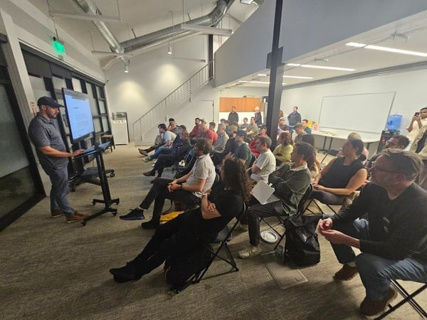 A photograph capturing a business event or presentation indoors, where a man stands giving a talk using a large monitor to a sizable audience seated in rows.