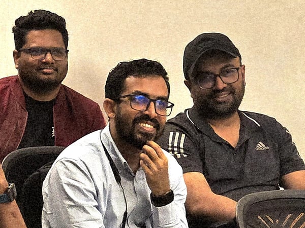 A close-up photograph shows three men, all wearing glasses, seated indoors and smiling while seemingly attending a presentation or meeting.