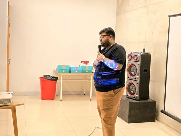 A man wearing glasses and a black t-shirt is giving a presentation indoors, holding a microphone. He stands next to a large stacked speaker system, with a projection screen and a table holding boxes visible in the background.