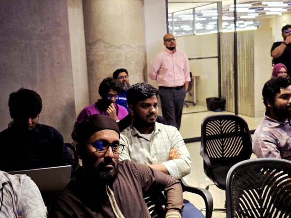 A candid indoor photograph shows a group of people, primarily men, seated and attending an event or presentation in a dimly lit room, with several individuals looking towards an unseen focal point.