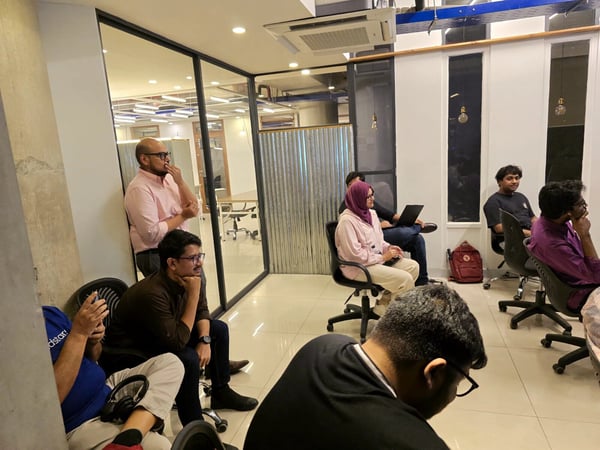 A candid indoor photograph captures a diverse group of people seated and standing in an office or co-working space, attentively facing an unseen focal point, suggesting they are attending a presentation or meeting.