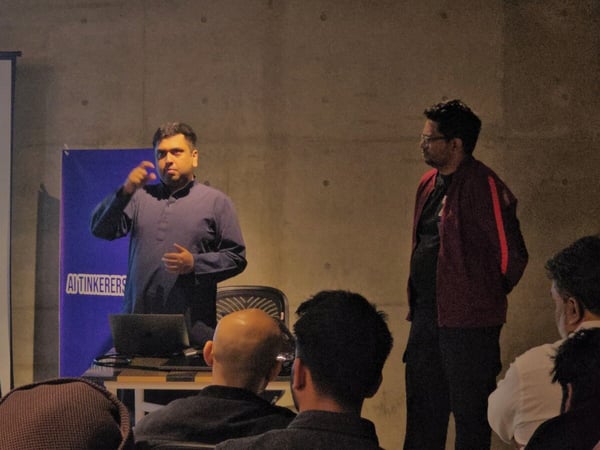 A photograph capturing an indoor technical presentation where a speaker, dressed in dark attire, stands gesturing beside a projected screen displaying text, while an audience watches from the foreground.