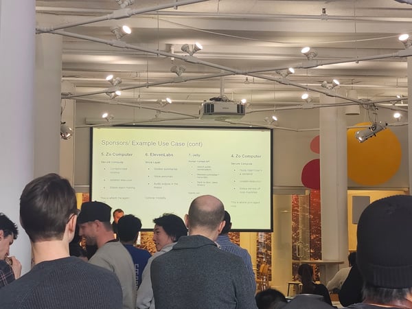 A photograph showing an indoor presentation where a crowd of people is watching a projector screen displaying information about tech sponsors like Zo Computer and ElevenLabs.