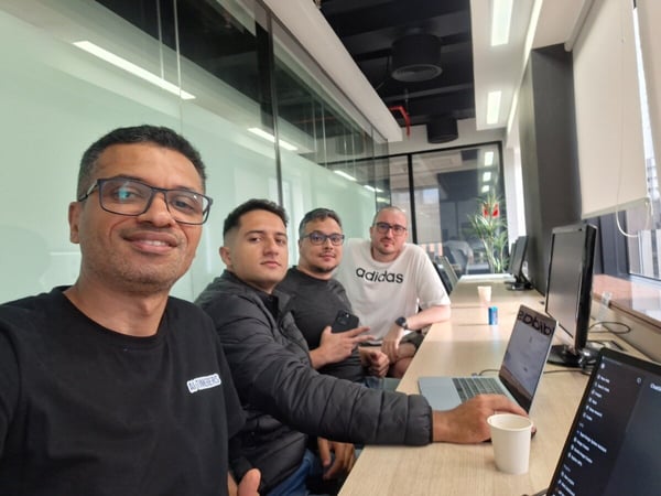 A group of four men are posing for a selfie while sitting at a long wooden desk in a modern office or coworking space.