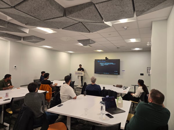 A man stands at a podium giving a presentation to a small audience seated at tables in a modern office or meeting room.