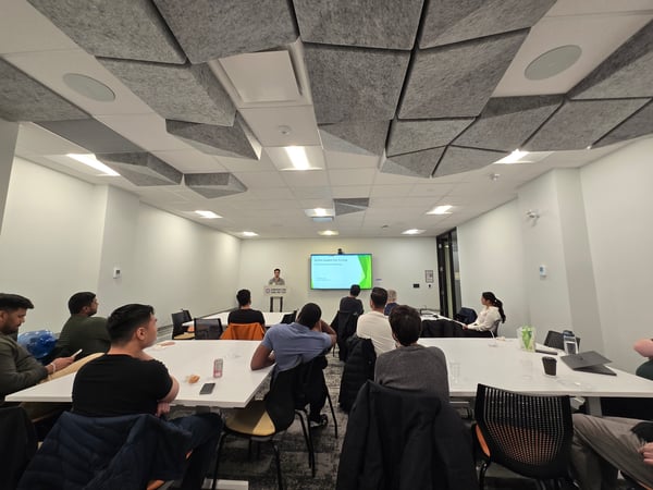 A man stands at a podium delivering a presentation to an audience seated at tables in a modern meeting room with acoustic ceiling tiles.
