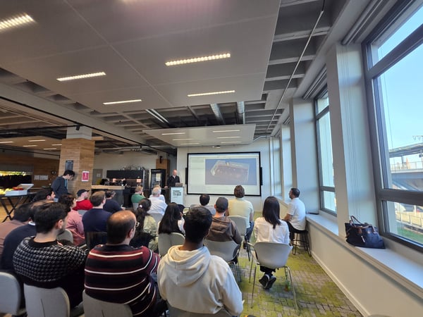 A group of people are seated in a modern office space, attending a presentation where a speaker stands next to a large screen displaying a 3D model.