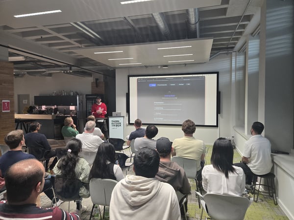 A group of people are seated in a modern office environment, watching a presentation on a large screen while a man in a red sweatshirt stands at the front.