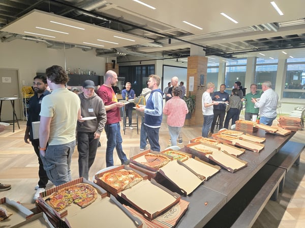 A group of people are gathered in a modern office or event space, socializing and eating pizza from several open boxes laid out on a long table.
