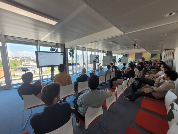 A group of people sitting in a bright, modern conference room with large windows, attending a presentation shown on a large digital screen.