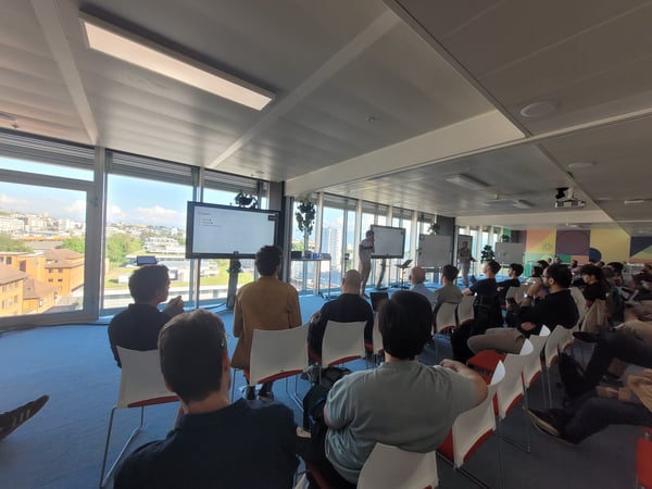 An indoor photograph of a presentation or workshop taking place in a modern office space with large windows overlooking a city.
