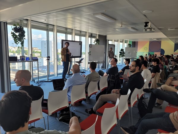 A man is giving a presentation to a seated audience in a modern office space with large windows overlooking a city.
