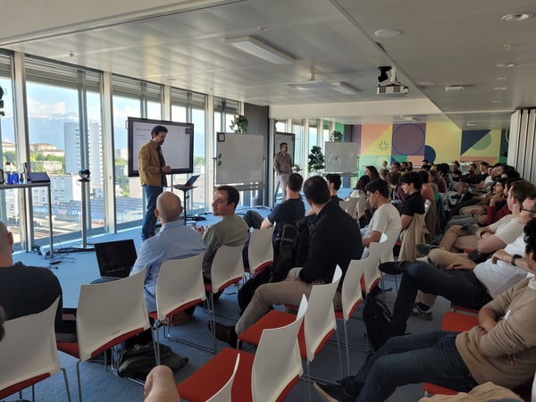 A man stands at the front of a modern conference room giving a presentation to a seated audience, with large windows showing a city view in the background.