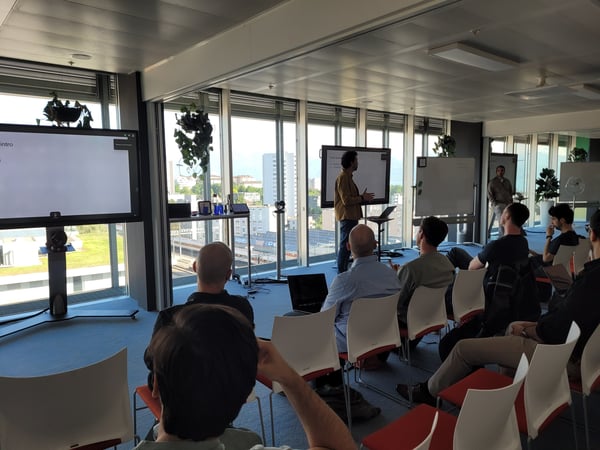 A group of people attending a presentation in a modern office with large windows overlooking a city view. A presenter stands at the front near digital screens and whiteboards while the audience sits in red-seated chairs.