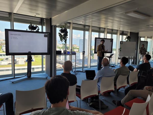 A man stands giving a presentation to an audience in a modern office with large windows overlooking a city and distant mountains.