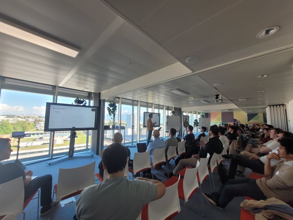 A large group of people are seated in a modern office space with floor-to-ceiling windows, attending a presentation.