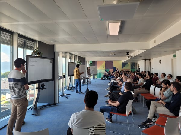 A group of people attending a presentation in a modern, well-lit office space with large windows and digital screens.