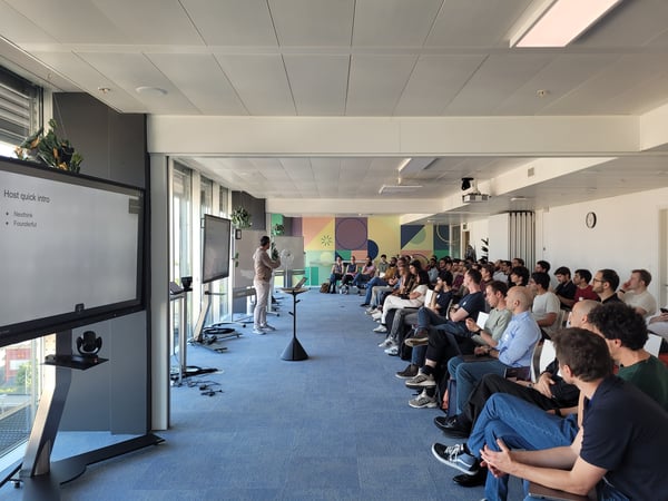 A man delivers a presentation to a large, seated audience in a modern, brightly lit office space.