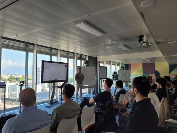 A man stands at the front of a modern office room giving a presentation to a seated audience, with large windows in the background showing a city and distant mountains.