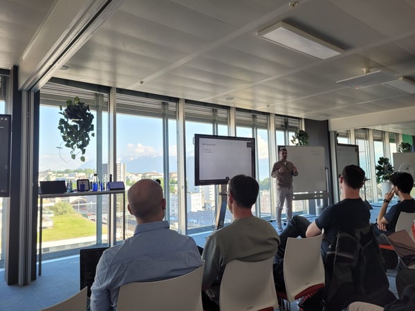 A man is giving a presentation to a small audience in a modern office space with large windows overlooking a city and distant mountains.